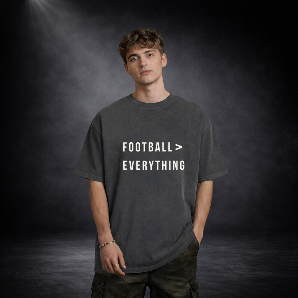 Männliches Model trägt ein dunkelgraues Oversized T-Shirt mit weißem Schriftzug „Football > Everything“, lässige Haltung mit Hand in der Tasche, vor dunklem Studio-Hintergrund mit weichem Spotlight-Licht