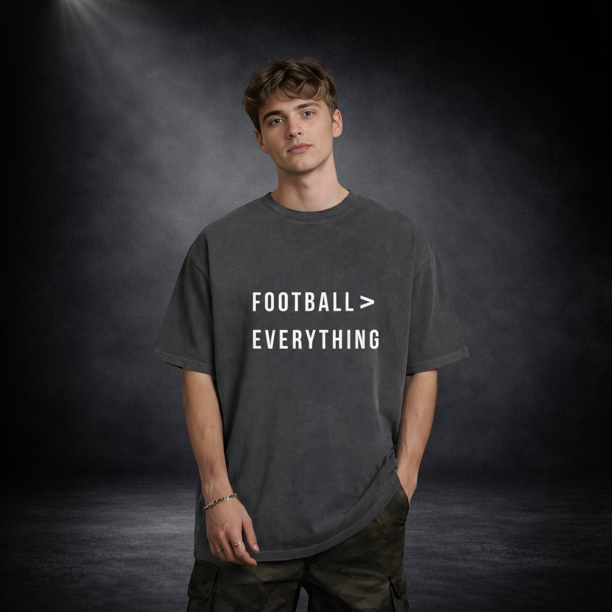 Männliches Model trägt ein dunkelgraues Oversized T-Shirt mit weißem Schriftzug „Football > Everything“, lässige Haltung mit Hand in der Tasche, vor dunklem Studio-Hintergrund mit weichem Spotlight-Licht