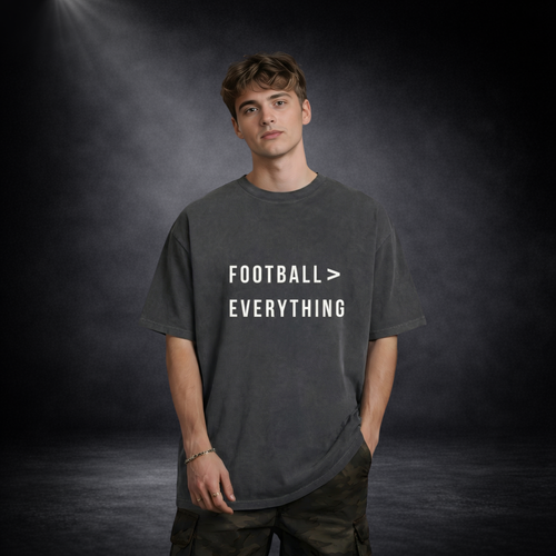 Männliches Model trägt ein dunkelgraues Oversized T-Shirt mit weißem Schriftzug „Football > Everything“, lässige Haltung mit Hand in der Tasche, vor dunklem Studio-Hintergrund mit weichem Spotlight-Licht