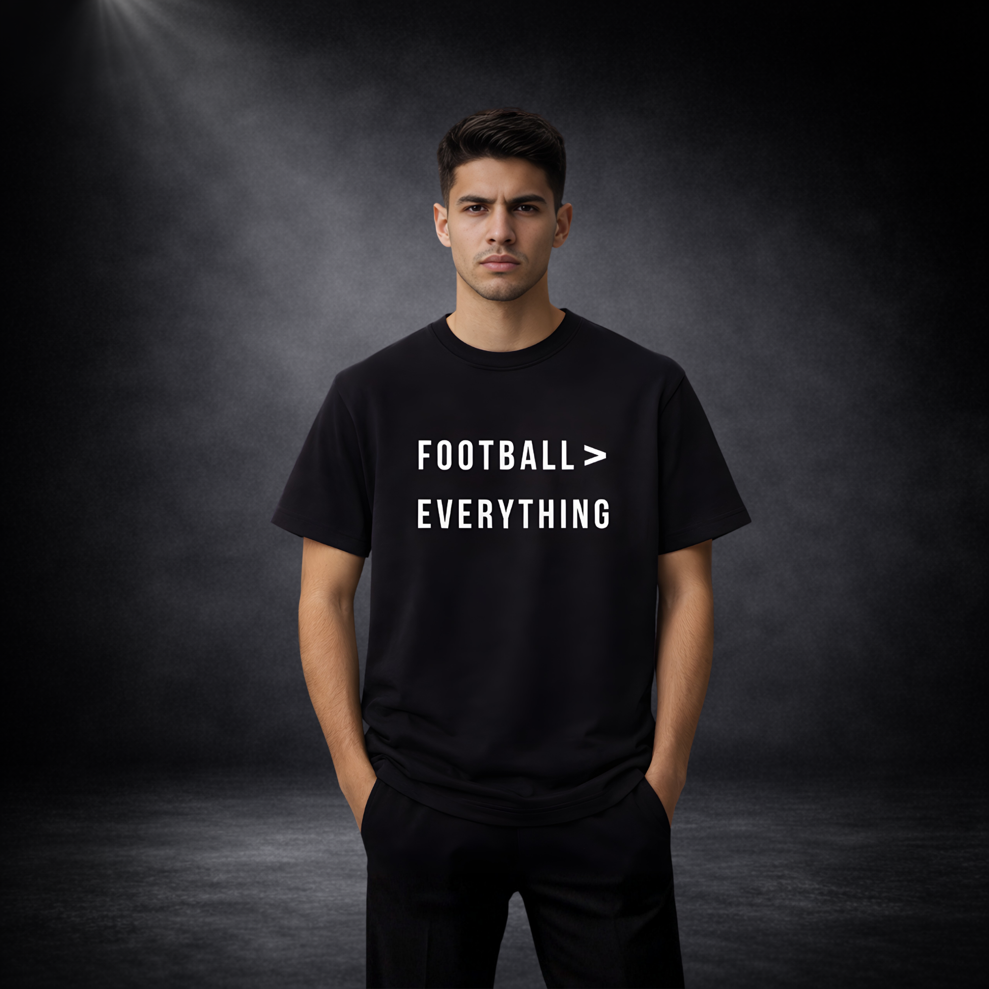 Männliches Model trägt schwarzes T-Shirt vor dunklem Studio-Hintergrund, Aufdruck „Football > Everything“ in Weiß auf der Vorderseite, lässige Pose mit Händen in den Taschen