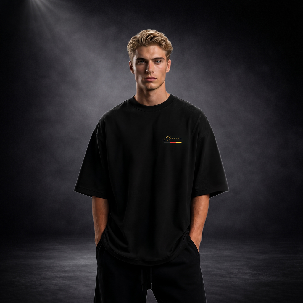 Blonder junger Mann trägt ein schwarzes Oversized T-Shirt von CERTANU in einer professionellen Studioaufnahme mit dunklem Premium-Hintergrund und weicher Spotlight-Beleuchtung. Das minimalistische Design zeigt ein kleines goldfarbenes CERTANU-Logo auf der linken Brust mit dezenten Akzenten in Schwarz, Rot und Gold. Das locker geschnittene Shirt vermittelt einen modernen Streetwear- und Fashion-Look. Der Fokus liegt auf der hochwertigen Passform, dem eleganten Oversized-Schnitt und dem schlichten Premium-Des