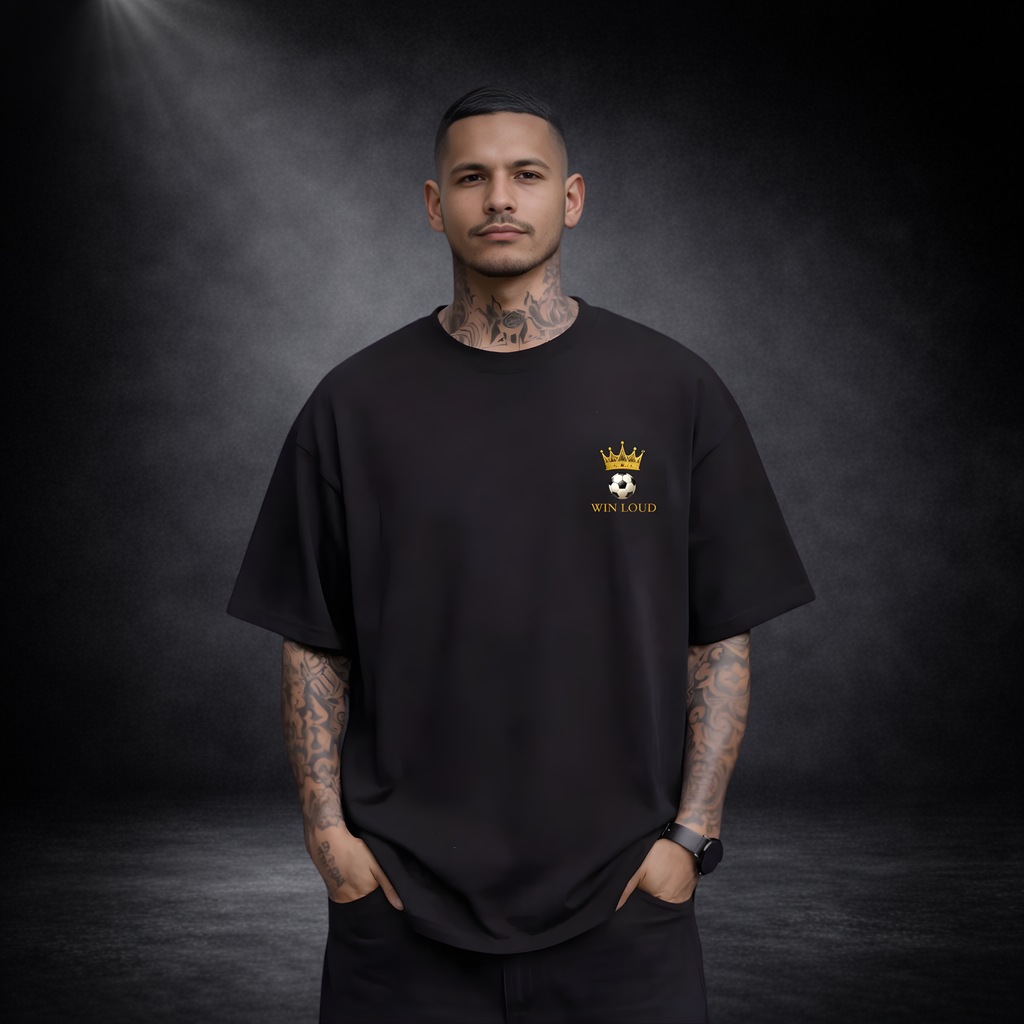 Mann mit Tattoos trägt ein schwarzes Oversized T-Shirt mit kleinem goldenen Kronen-Fußball-Logo und „WIN LOUD“ Schriftzug auf der Brust vor dunklem Studio-Hintergrund