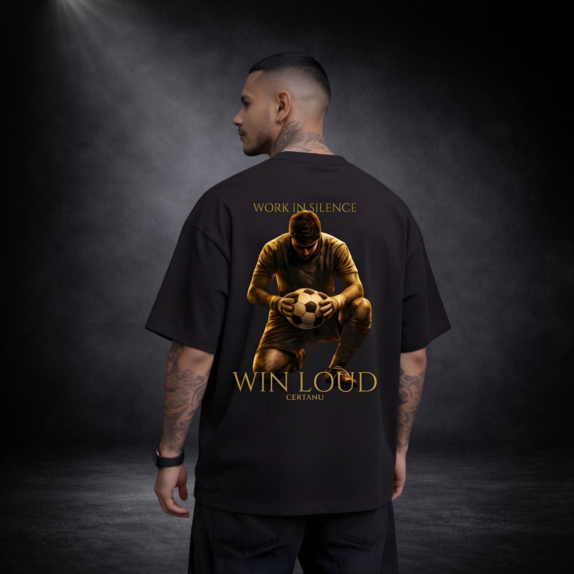 Rückansicht eines Mannes mit Tattoos in schwarzem Oversized T-Shirt, großes goldenes Fußball-Motiv mit kniendem Spieler, Text „WORK IN SILENCE“ und „WIN LOUD CERTANU“ auf dem Rücken, dunkler Studio-Hintergrund
