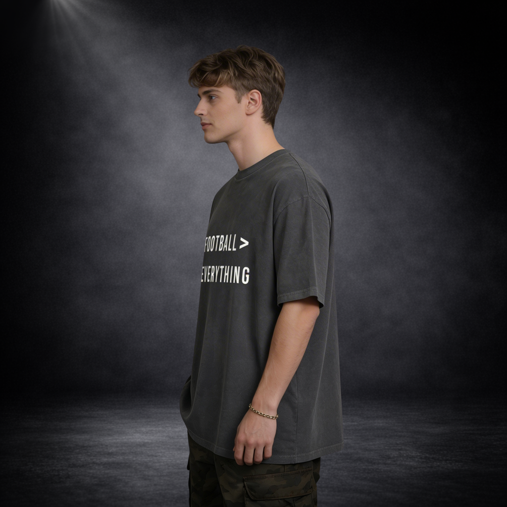 Seitliche Ansicht eines männlichen Models in dunkelgrauem Oversized T-Shirt mit „Football > Everything“-Print, lässige Haltung vor dunklem Studio-Hintergrund mit weichem Licht und leichter Nebelwirkung