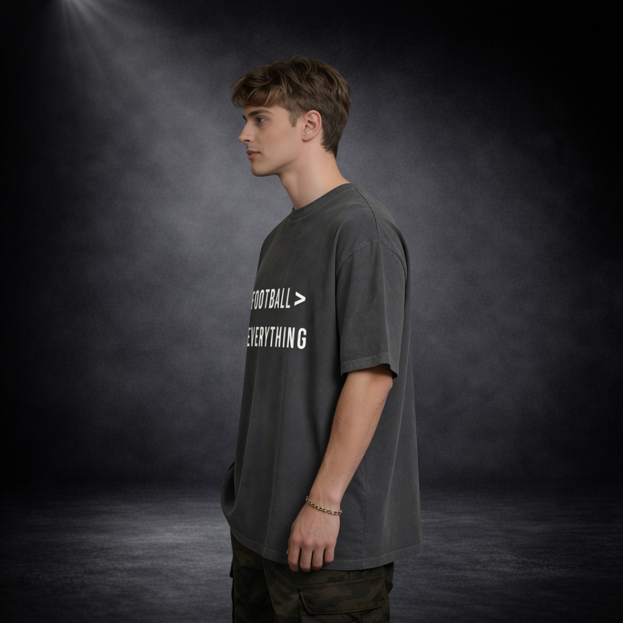 Seitliche Ansicht eines männlichen Models in dunkelgrauem Oversized T-Shirt mit „Football > Everything“-Print, lässige Haltung vor dunklem Studio-Hintergrund mit weichem Licht und leichter Nebelwirkung