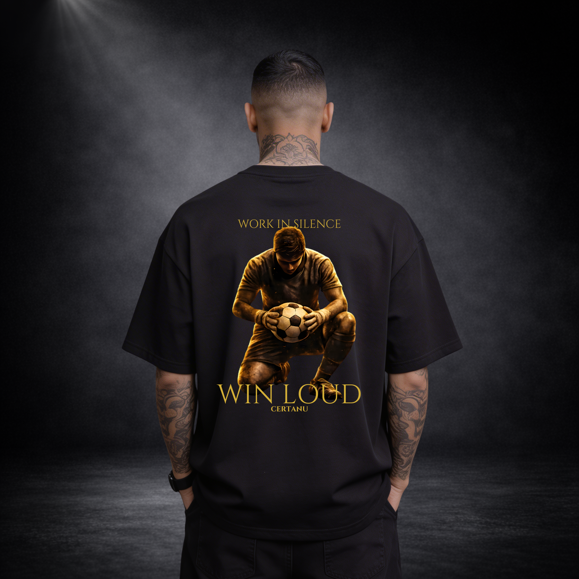 Rückansicht eines Mannes mit Nacken- und Arm-Tattoos im schwarzen Oversized T-Shirt, großes goldenes Motiv eines knienden Fußballspielers mit Ball, darüber „WORK IN SILENCE“ und darunter „WIN LOUD CERTANU“, vor dunklem Studio-Hintergrund