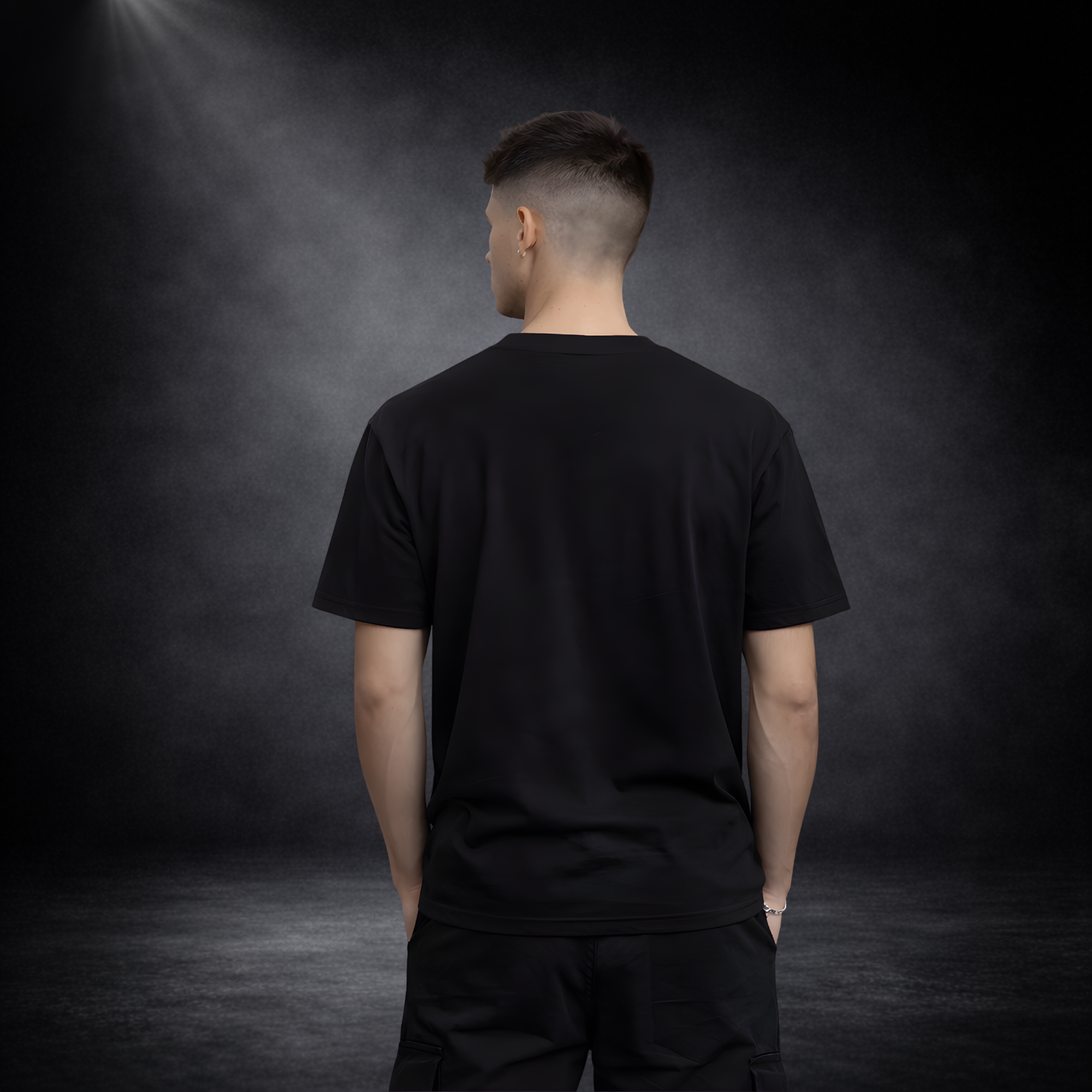 Rückansicht eines jungen Mannes in schwarzem T-Shirt ohne Aufdruck, Hände in den Taschen, vor dunklem Studio-Hintergrund mit Spotlight