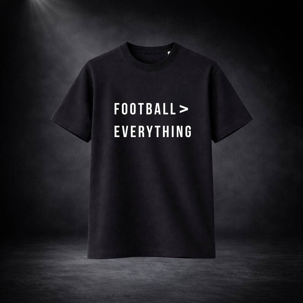 Schwarzes T-Shirt vor dunklem Studio-Hintergrund, frontal dargestellt, mit weißem Aufdruck „FOOTBALL > EVERYTHING“ in klarer, moderner Schrift
