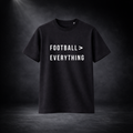 Schwarzes T-Shirt vor dunklem Studio-Hintergrund, frontal dargestellt, mit weißem Aufdruck „FOOTBALL > EVERYTHING“ in klarer, moderner Schrift