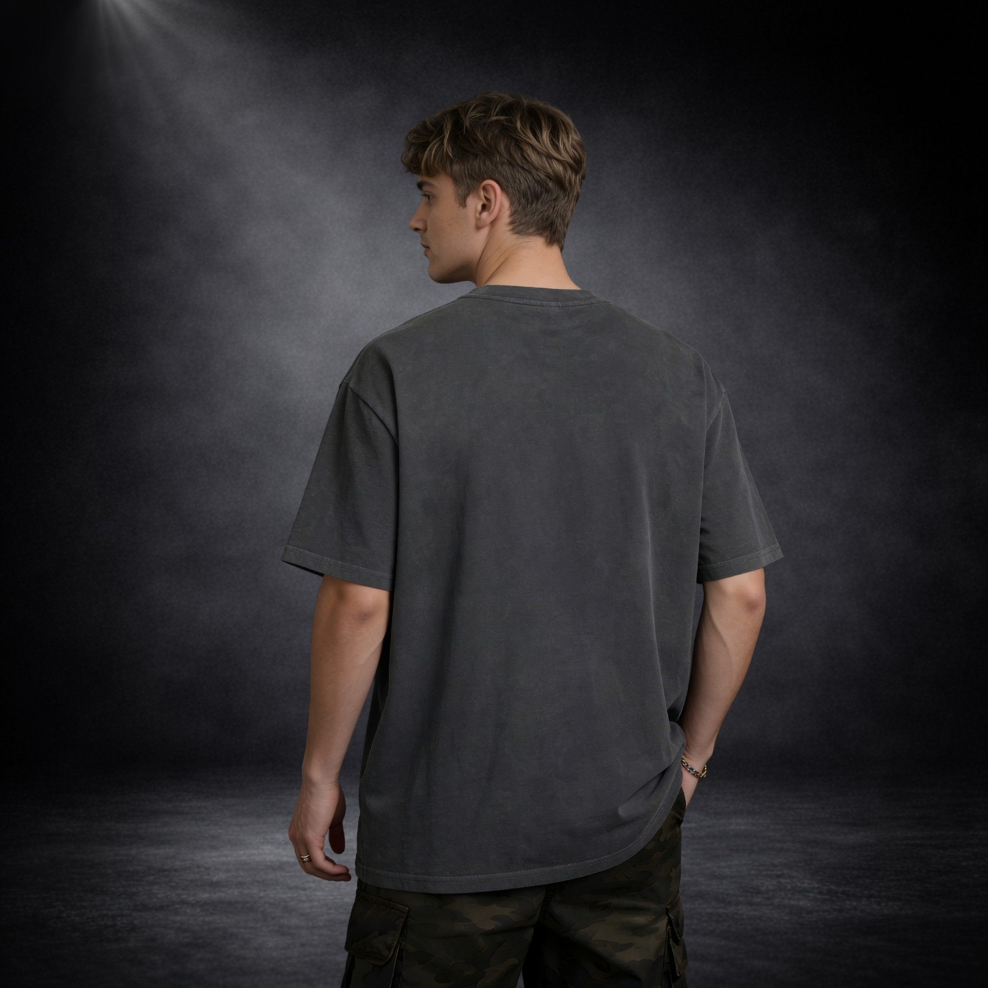 Rückansicht eines männlichen Models in dunkelgrauem Oversized T-Shirt ohne Aufdruck, lockere Passform, vor dunklem Studio-Hintergrund mit weichem Spotlight und leichter Nebelstimmung