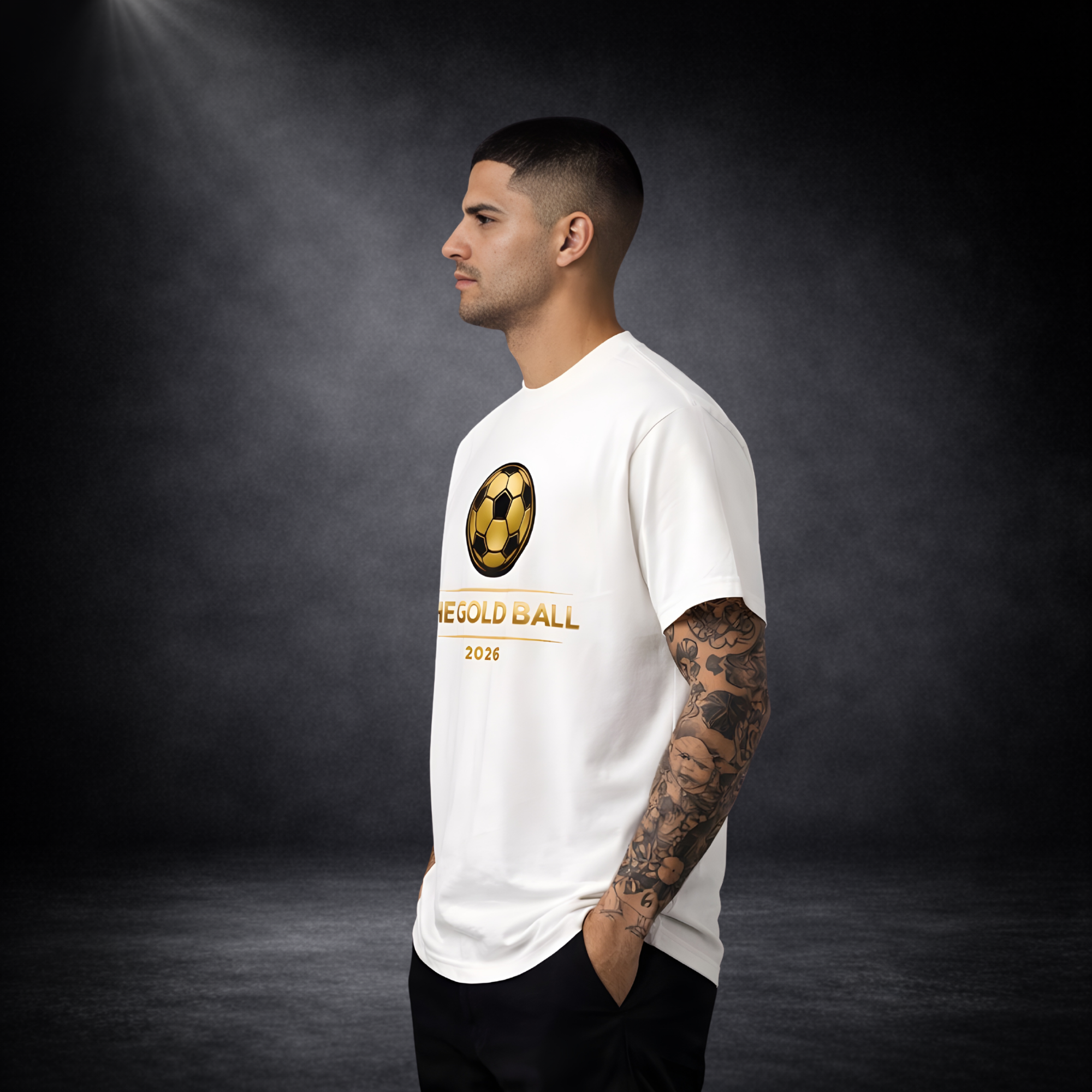 Seitliche Ansicht eines jungen Mannes mit tätowiertem Arm in einem weißen T-Shirt mit goldenem Fußball-Logo und Schriftzug „THE GOLD BALL 2026“, vor dunklem Studio-Hintergrund mit weichem Spotlight