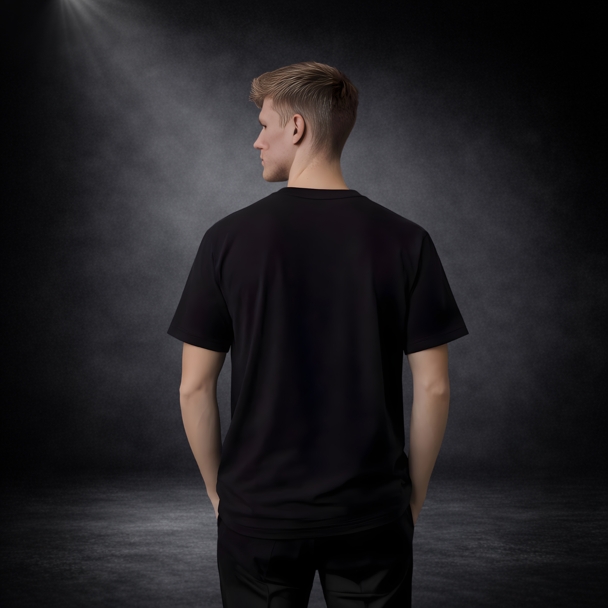Rückansicht eines jungen Mannes in einem schlichten schwarzen T-Shirt vor dunklem Studio-Hintergrund, ohne Aufdruck; weiches Spotlight von oben links, Hände in den Hosentaschen