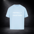 Hellblaues Oversized T-Shirt mit weißem Schriftzug „FOOTBALL > EVERYTHING“ auf der Vorderseite, zentriert dargestellt vor dunklem, leicht nebligem Studio-Hintergrund mit Spotlight-Beleuchtung
