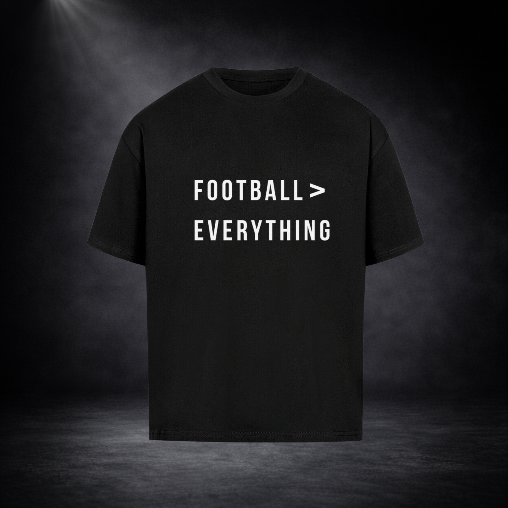 Schwarzes Oversized T-Shirt mit weißem Schriftzug „FOOTBALL > EVERYTHING“ auf der Vorderseite, mittig platziert, vor dunklem Studio-Hintergrund mit dezentem Lichtspot und leichtem Nebeleffekt