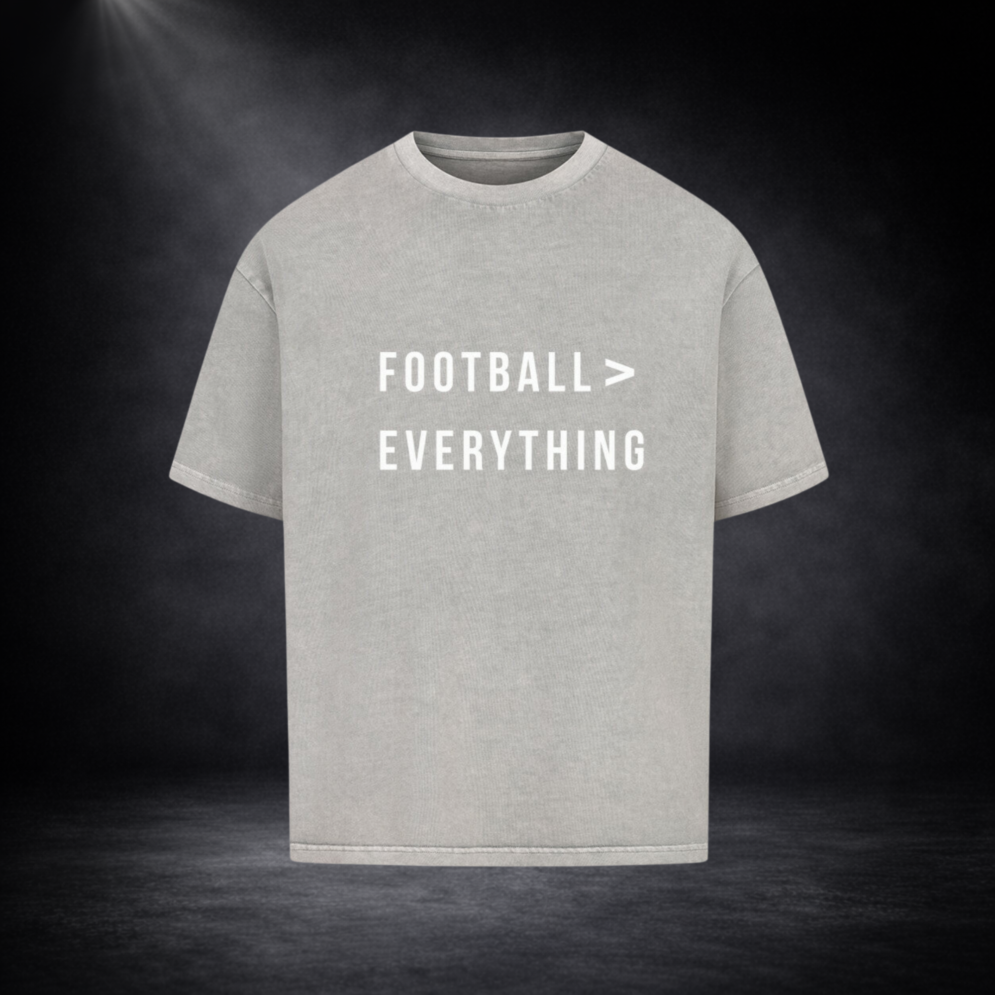 Graues Oversized T-Shirt mit weißem Schriftzug „FOOTBALL > EVERYTHING“ auf der Vorderseite, zentriert platziert vor dunklem Studio-Hintergrund mit sanftem Spotlight und leichtem Nebeleffekt