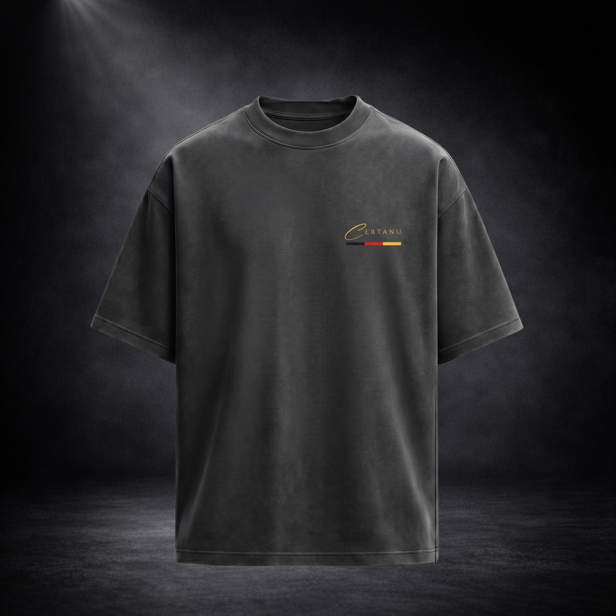 Vorderansicht eines dunkelgrauen Oversized CERTANU T-Shirts vor einem eleganten dunklen Studiohintergrund mit dezenter Spotlight-Beleuchtung. Das minimalistische Design zeigt auf der linken Brust ein hochwertiges CERTANU-Logo in Gold mit einer feinen Schwarz-Rot-Gold-Linie als Anspielung auf die deutschen Nationalfarben. Das Premium-Shirt besitzt eine moderne Oversized-Passform mit überschnittenen Schultern, breiten Ärmeln und hochwertiger Stoffstruktur. Die schlichte Streetwear-Optik kombiniert Luxus, Fußb
