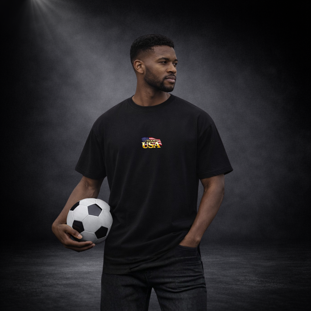 Mann trägt schwarzes T-Shirt mit kleinem „Team USA“-Logo auf der Brust und hält einen Fußball, vor dunklem Studio-Hintergrund