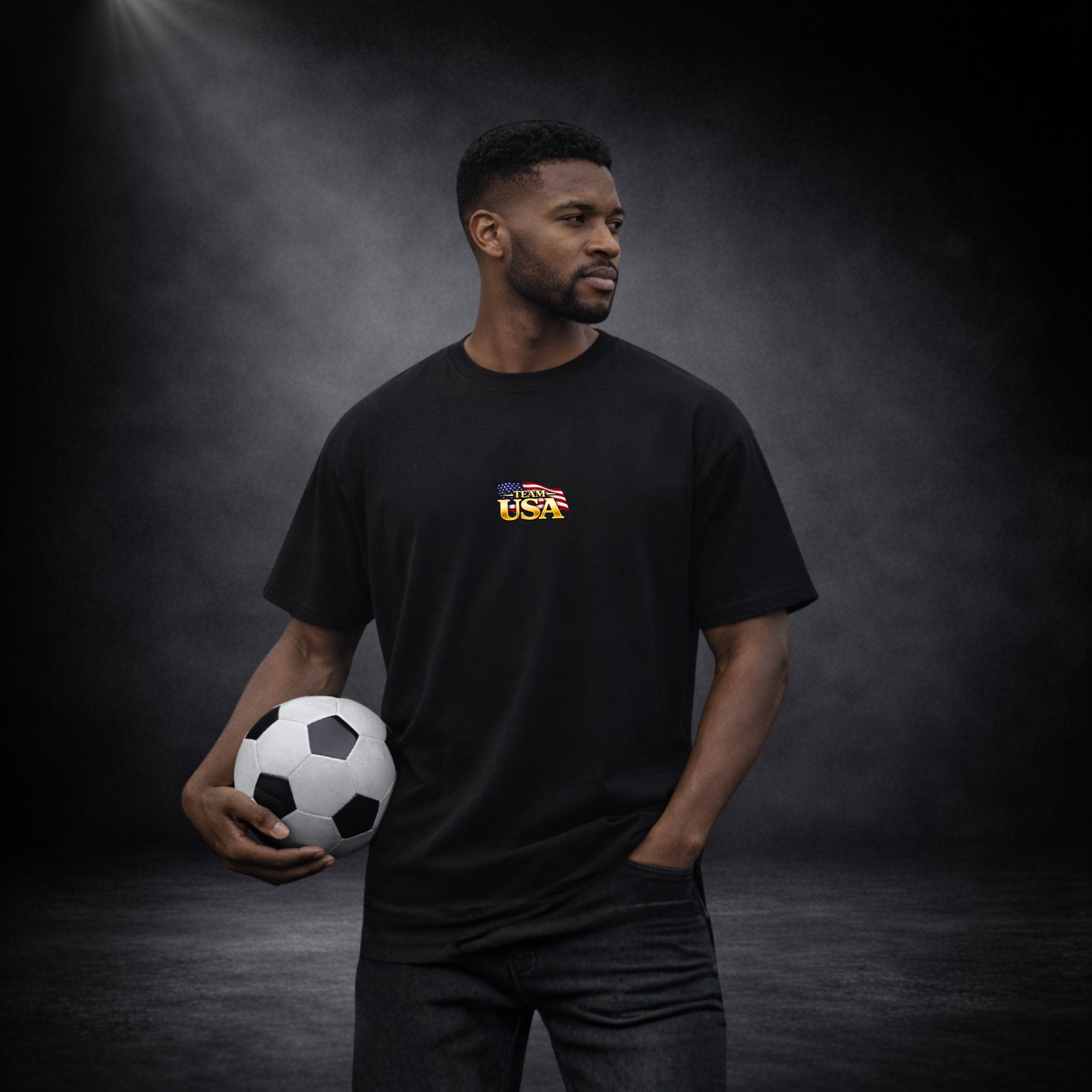 Mann trägt schwarzes T-Shirt mit kleinem „Team USA“-Logo auf der Brust und hält einen Fußball, vor dunklem Studio-Hintergrund