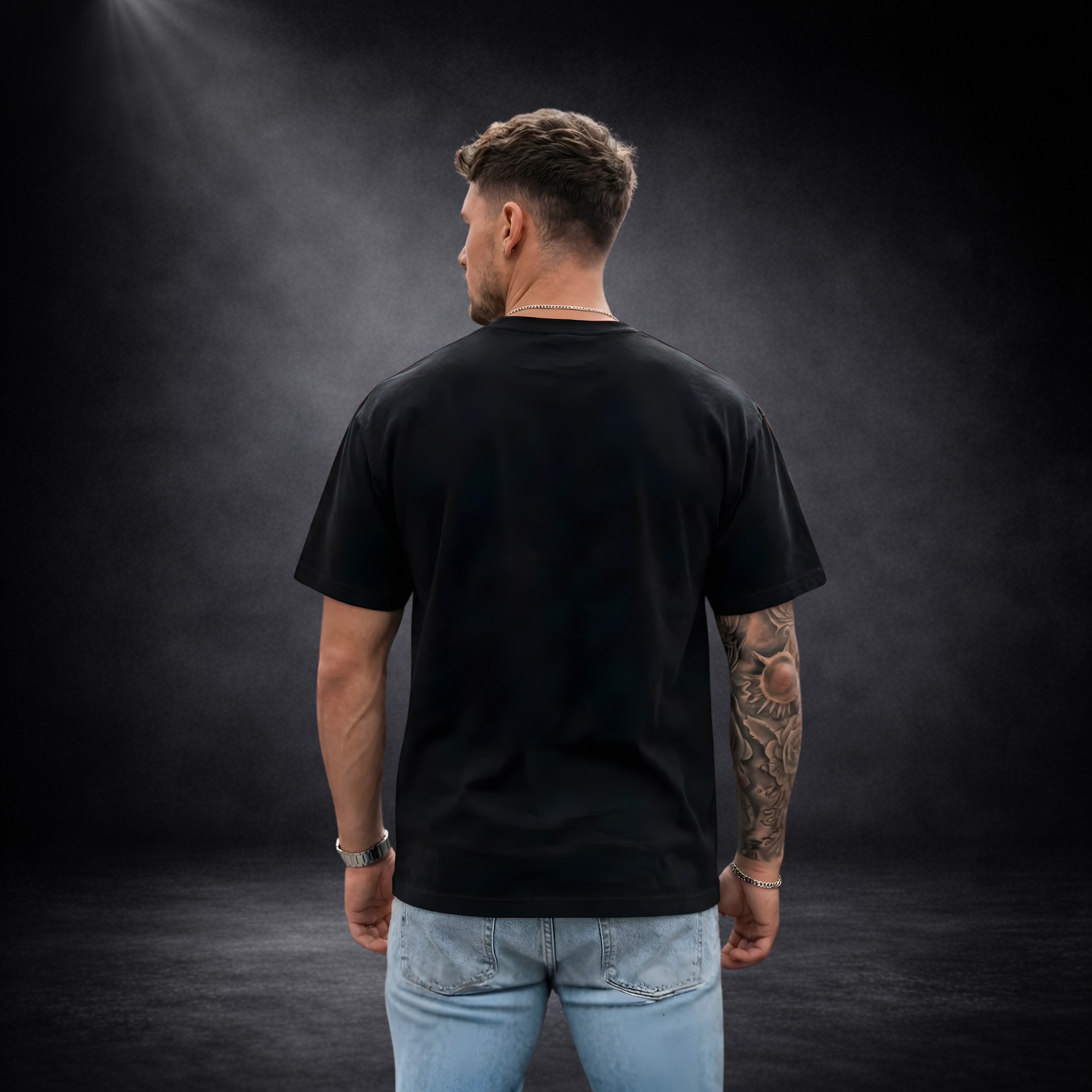 Männliches Model von hinten in schwarzem T-Shirt ohne Aufdruck, kombiniert mit Jeans, vor dunklem Studio-Hintergrund