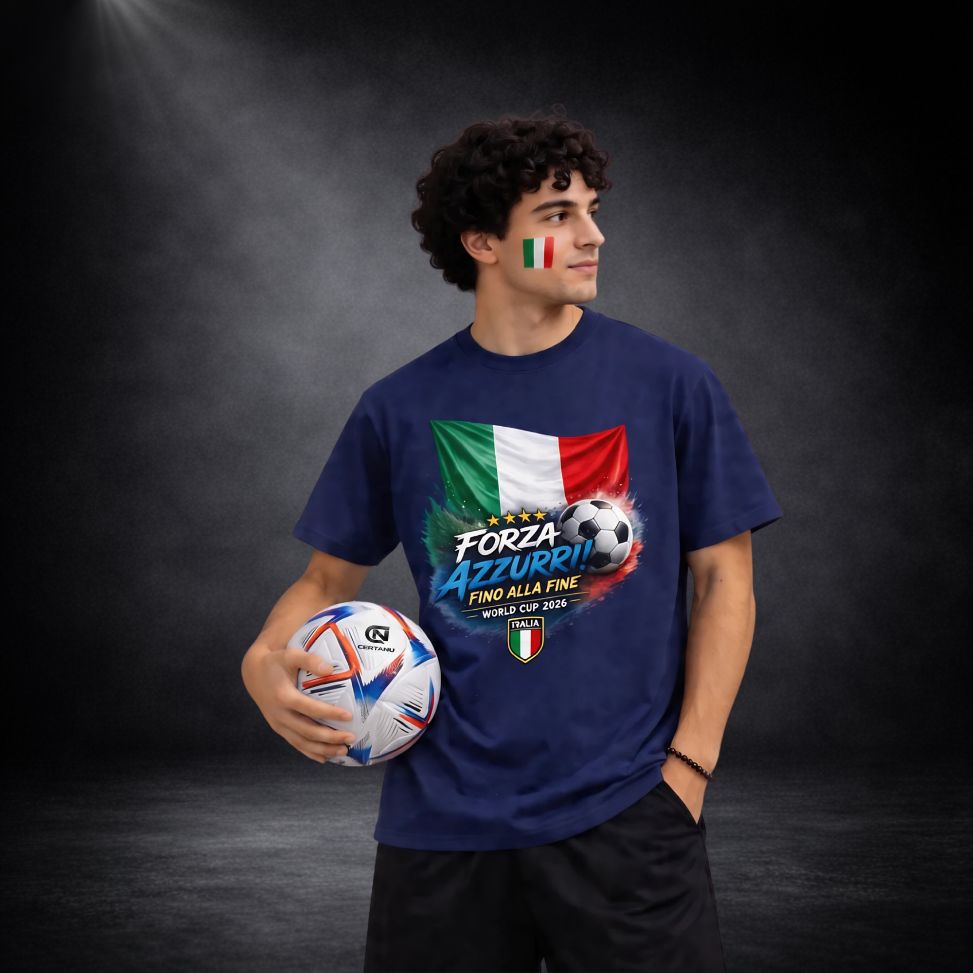 Junger Mann mit Italien-Flagge im Gesicht trägt blaues „Forza Azzurri – Fino Alla Fine World Cup 2026“ T-Shirt mit italienischer Flagge und Fußball, hält Ball und schaut zur Seite vor dunklem Studiohintergrund
