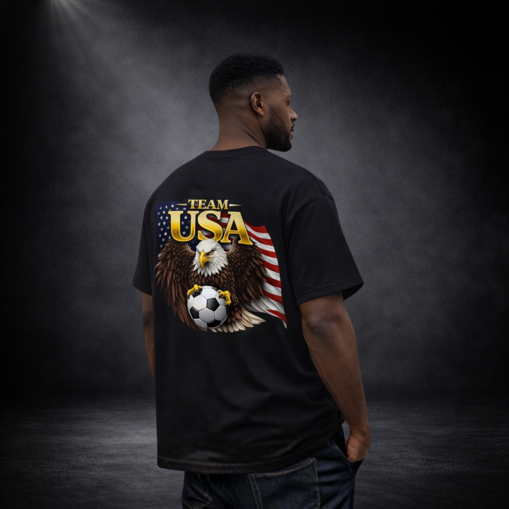 Rückansicht eines Mannes im oversized schwarzen T-Shirt mit großem „Team USA“-Print, Adler mit Fußball und US-Flagge vor dunklem Studio-Hintergrund