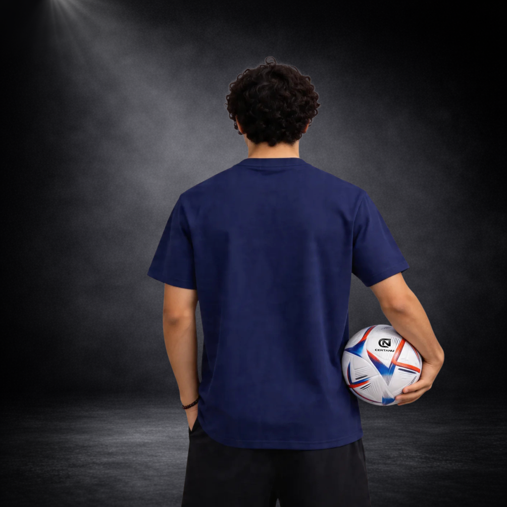 Rückansicht eines jungen Mannes mit blauem Italien Fußball T-Shirt ohne Druck, hält einen Fußball in der Hand, vor dunklem Studiohintergrund mit Spotlight-Beleuchtung