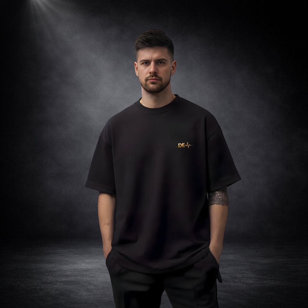 Mann in schwarzem Oversize-T-Shirt vor dunklem Studio-Hintergrund, mit kleinem goldenen Logo auf der Brust, minimalistisch und modern inszeniert