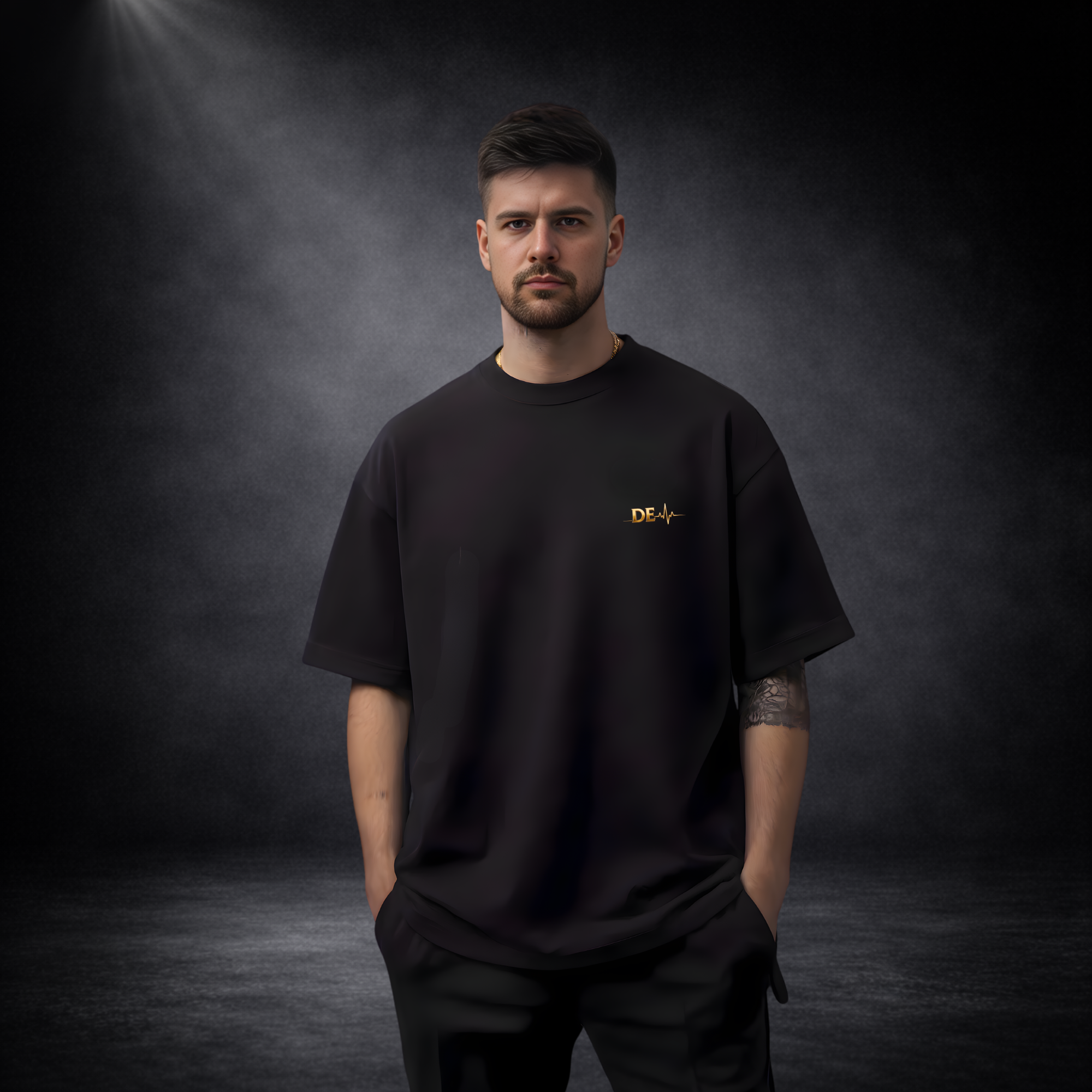 Mann in schwarzem Oversize-T-Shirt vor dunklem Studio-Hintergrund, mit kleinem goldenen Logo auf der Brust, minimalistisch und modern inszeniert