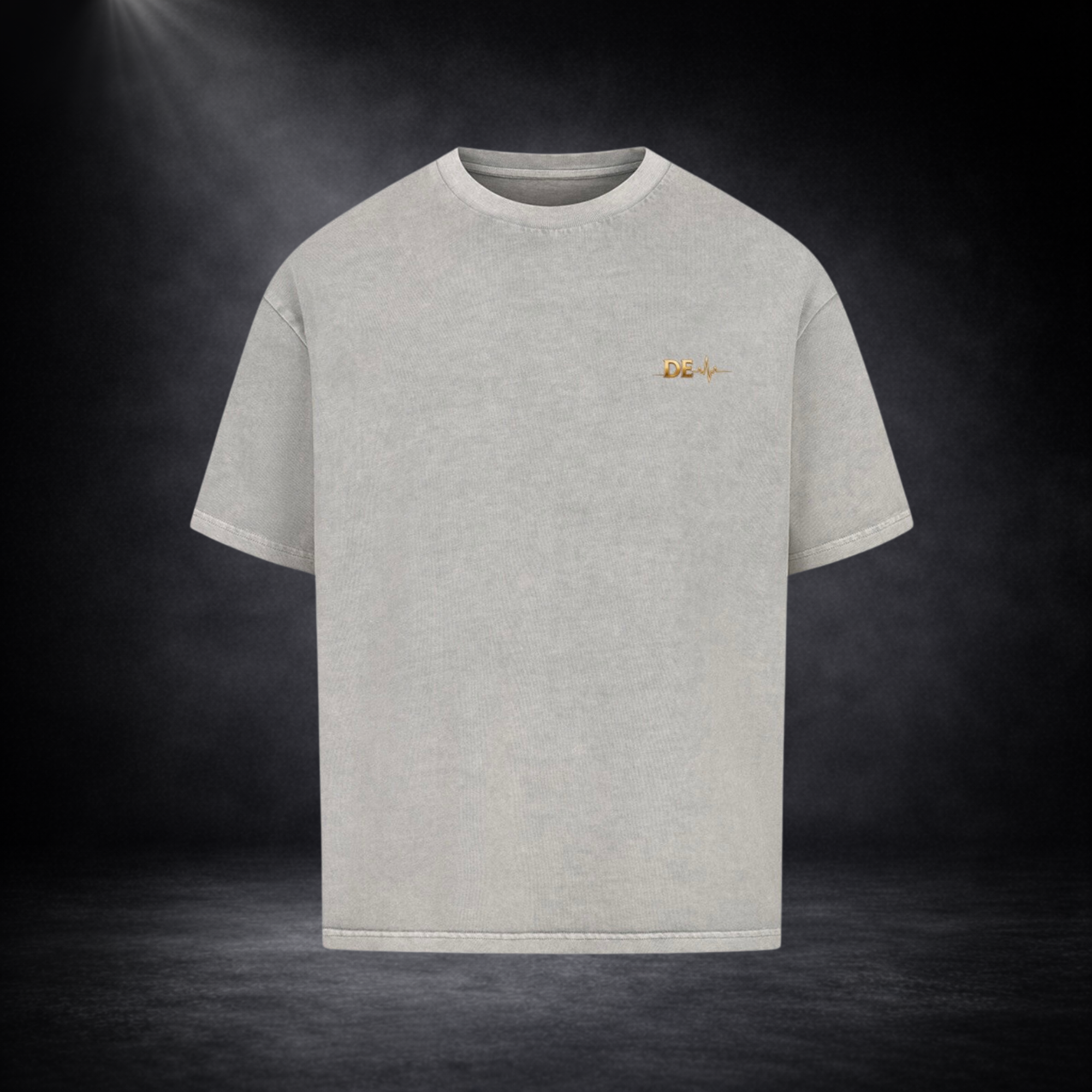 Hellgraues T-Shirt in Frontansicht vor dunklem Studio-Hintergrund, mit kleinem goldenen „DE“-Logo und Herzschlag-Linie auf der Brust in schlichtem Design