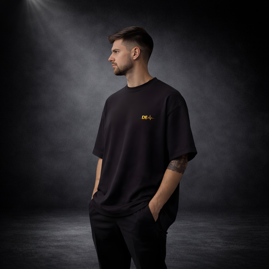Mann im Profil vor dunklem Studio-Hintergrund, trägt ein schwarzes Oversize-T-Shirt mit kleinem goldenem Brustlogo, minimalistischer und hochwertiger Look