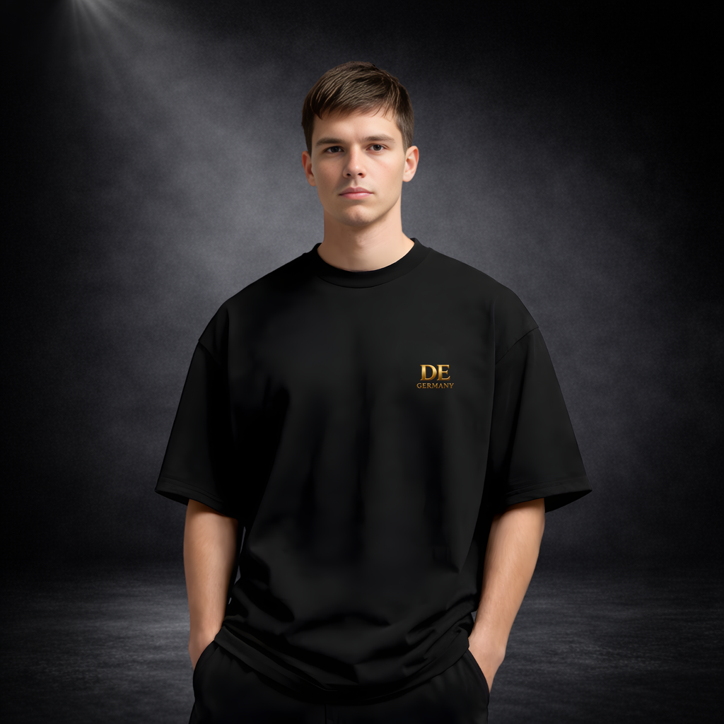 Mann trägt ein schwarzes T-Shirt vor dunklem Studio-Hintergrund, mit kleinem goldenen „DE Germany“-Logo auf der Brust in minimalistischem, edlem Design