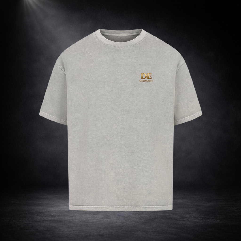 Hellgraues Oversized T-Shirt mit kleinem goldenen „DE Germany“-Print auf der Brust, dargestellt vor dunklem Studio-Hintergrund im cleanen Premium-Stil