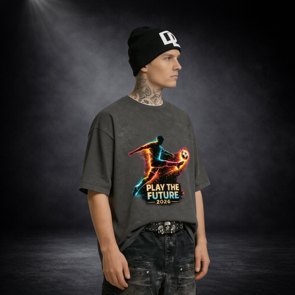 Männliches Model mit schwarzer Beanie und Tattoos trägt ein dunkelgraues T-Shirt mit leuchtendem Fußballspieler-Print und „Play the Future 2026“-Schriftzug vor dunklem Studio-Hintergrund