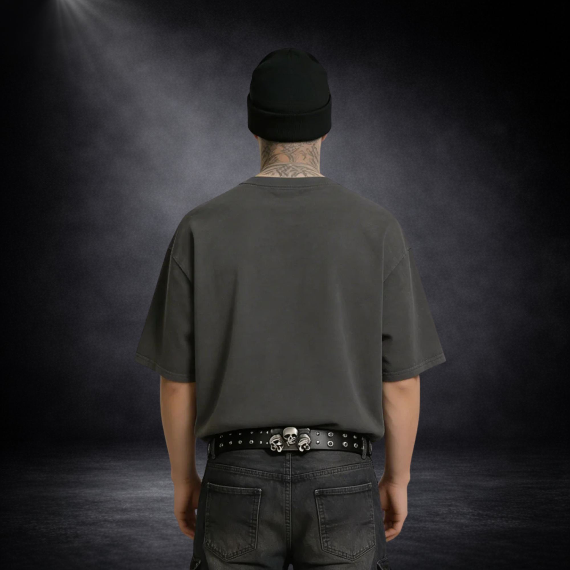 Rückansicht eines männlichen Models mit schwarzer Beanie und Nacken-Tattoo, das ein schlichtes dunkelgraues T-Shirt ohne Print trägt, kombiniert mit schwarzer Jeans und Gürtel vor dunklem Studio-Hintergrund