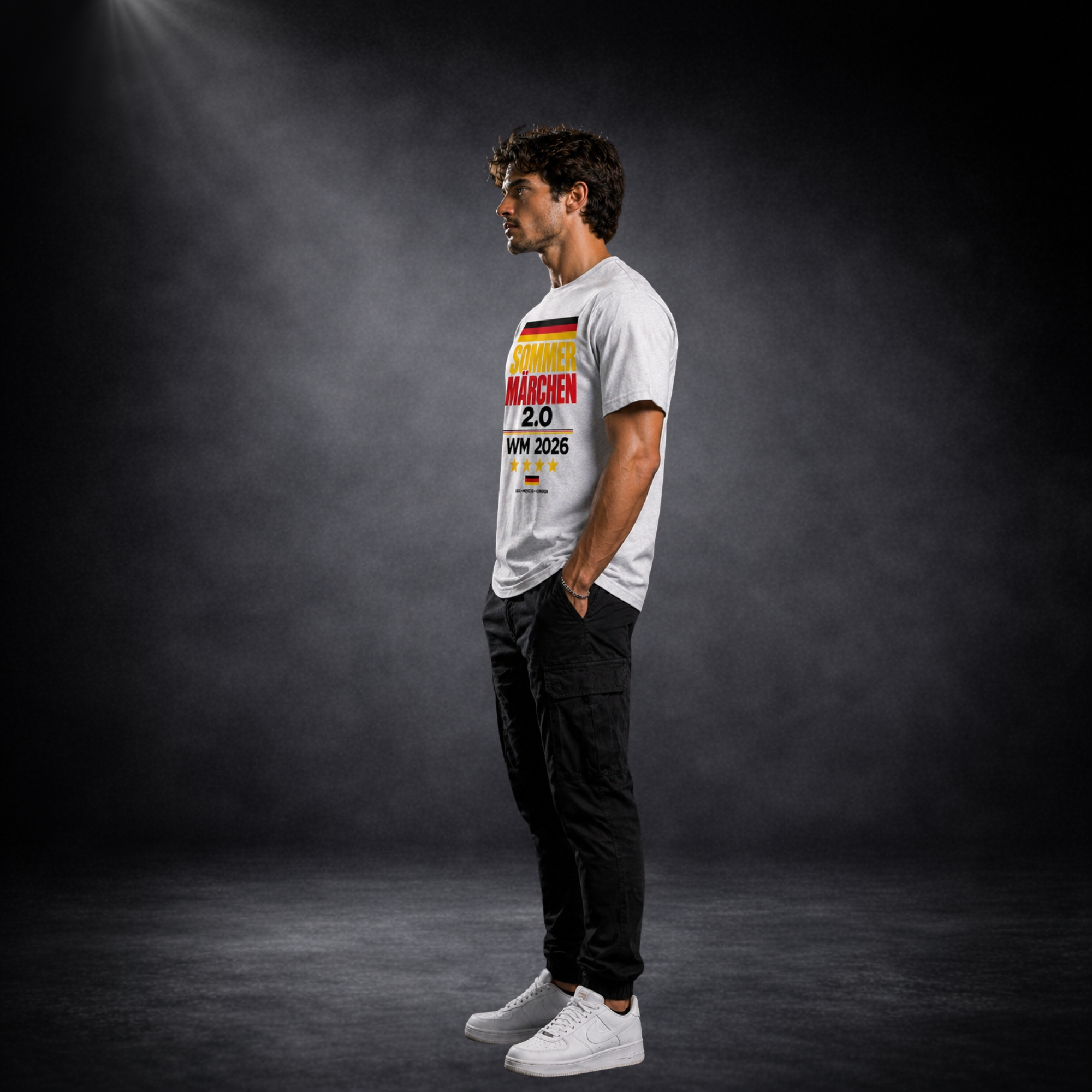 Seitliche Ansicht eines jungen Mannes im grauen Deutschland WM 2026 T-Shirt mit „Sommermärchen 2.0“ Print, sportlicher Streetwear Look, dunkler Studiohintergrund