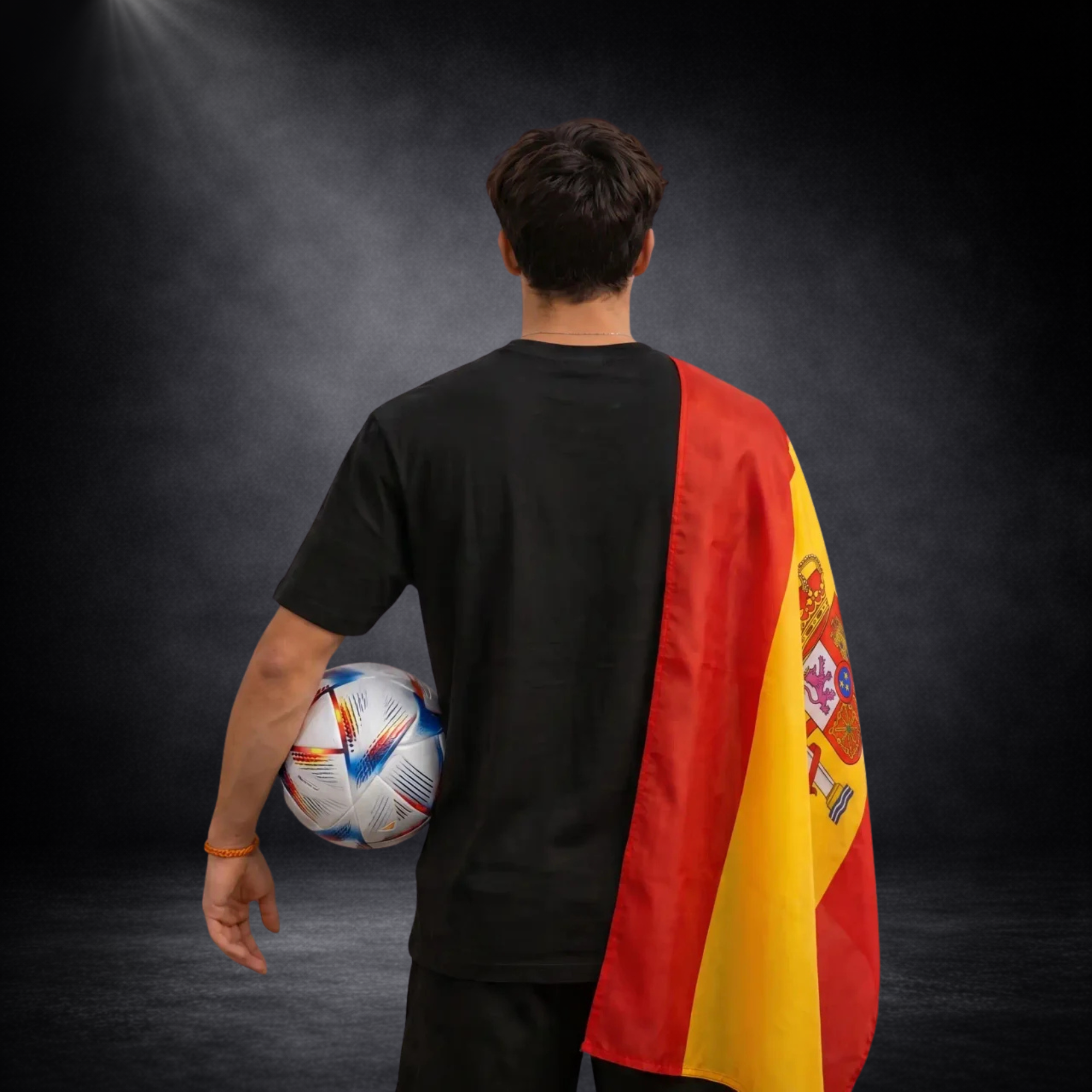 Rückansicht eines jungen Mannes mit Spanien-Flagge über der Schulter, schwarzes T-Shirt ohne Design, hält einen Fußball vor dunklem Studiohintergrund