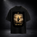 Schwarzes Oversized T-Shirt von CERTANU mit großem Frontprint vor dunklem Premium-Studiohintergrund. Das Design zeigt einen jungen Fußballspieler mit Ball, der auf ein hell erleuchtetes Stadion in der Ferne blickt. Warme Gold- und Lichttöne erzeugen eine emotionale Atmosphäre und symbolisieren den Traum vom Profifußball. Unter dem Motiv steht der Schriftzug „THE DREAM“ in goldener Typografie. Das hochwertige Fußball-Design vermittelt Motivation, Leidenschaft, Ehrgeiz und den Weg vom Nachwuchsspieler zur Leg