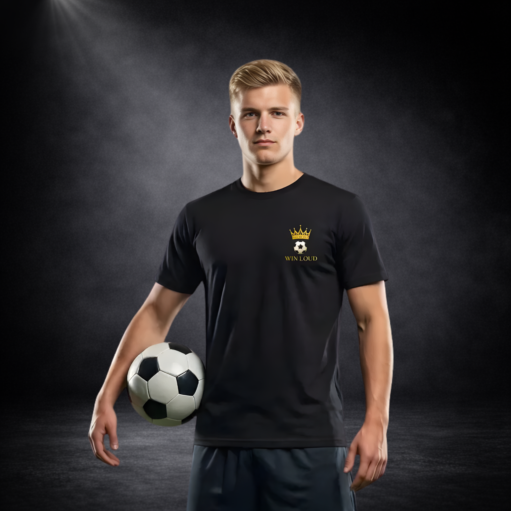 Junger Mann im dunklen Studio trägt ein schwarzes T-Shirt mit kleinem goldenen Fußball-Kronen-Logo auf der Brust und hält einen Fußball unter dem Arm