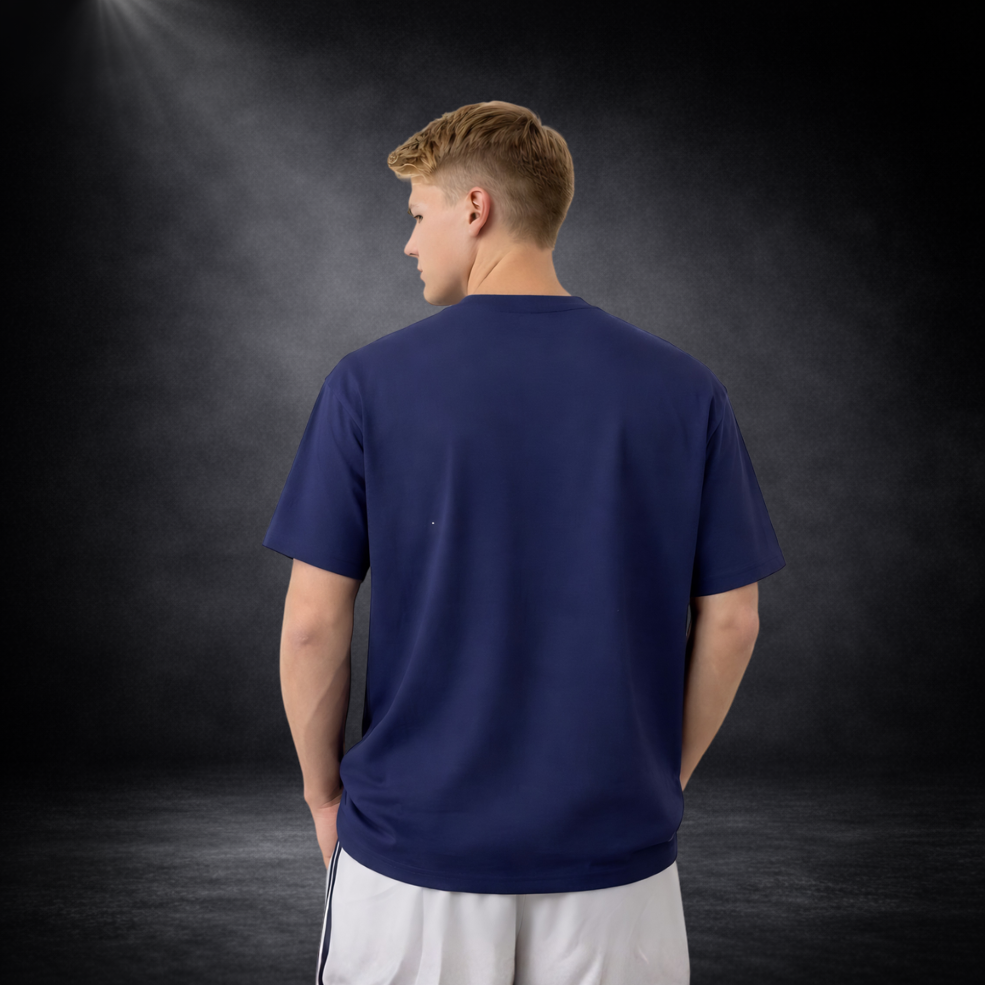 Rückansicht eines jungen Mannes mit kurzem blondem Haar, trägt ein schlichtes blaues T-Shirt ohne Druck, kombiniert mit weißen Shorts, vor dunklem Studio-Hintergrund